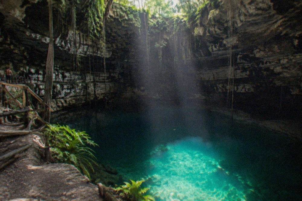 Sunlight beams entering a jungle cenote through an opening in the rock ceiling over clear blue-green water