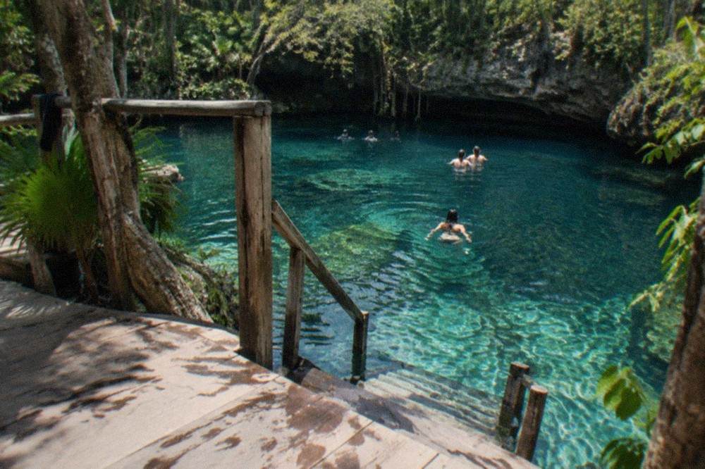 A wooden ladder and platform descending into a jungle sinkhole with pale blue water below