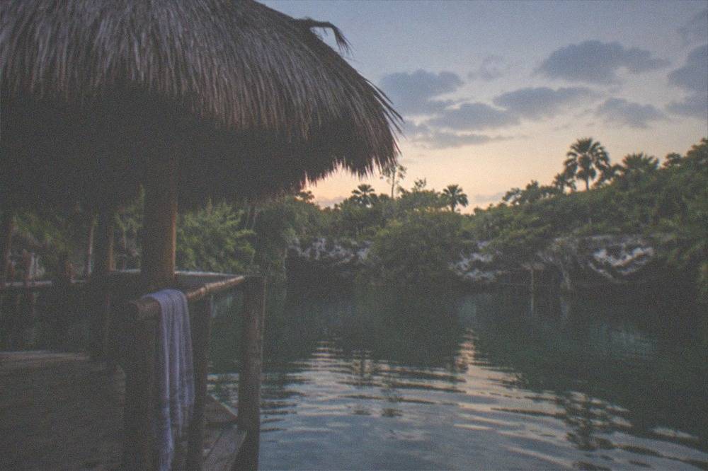 A thatched palapa shade structure beside a wooden dock over calm green cenote water