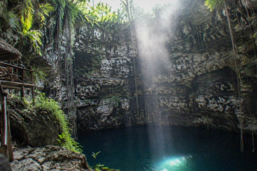 Sunlight streaming through a cenote opening onto still clear water and limestone formations