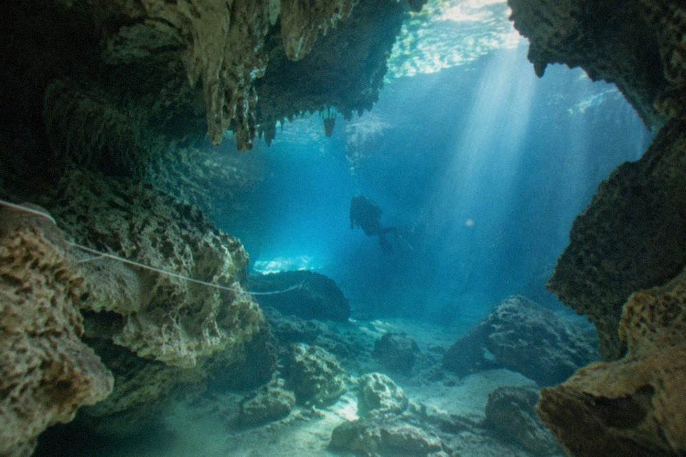 A diver's silhouette gliding through a sunlit limestone cavern passage with a guideline visible along the rock wall