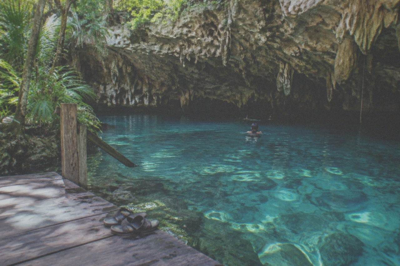 Clear turquoise water in a limestone cenote basin with stalactites hanging over a wooden swim platform