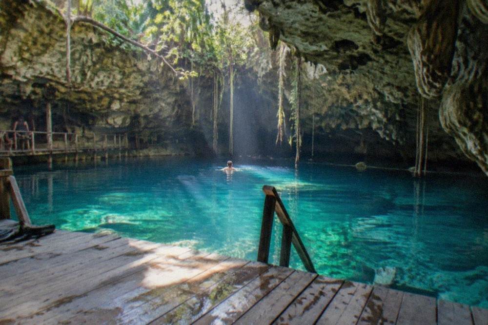 Sunlight beams entering turquoise water through openings in a limestone ceiling above a wooden swim platform