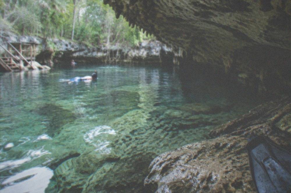 A snorkeler floating above pale limestone formations in clear shallow water beneath overhanging rock