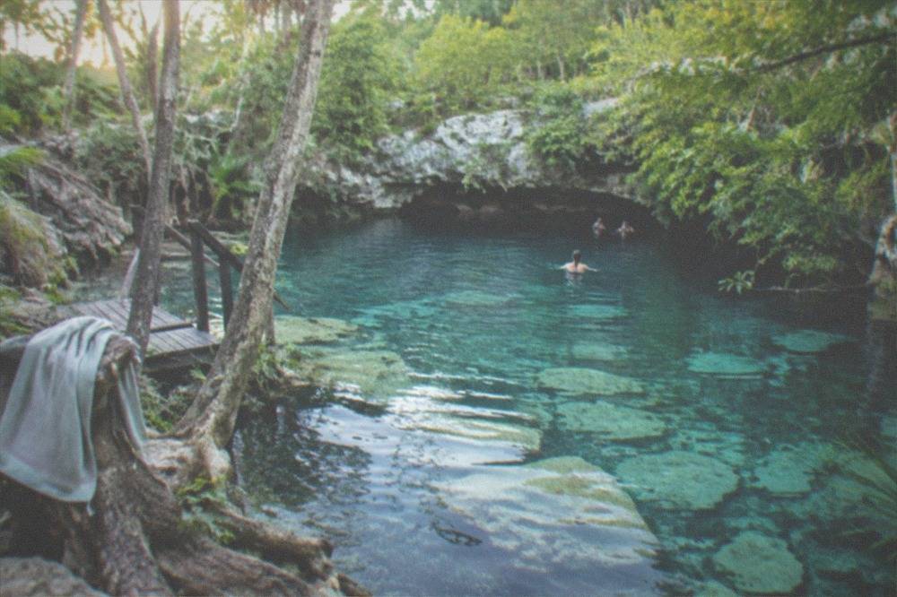 A round open cenote surrounded by jungle with pale sand and rock visible under transparent water