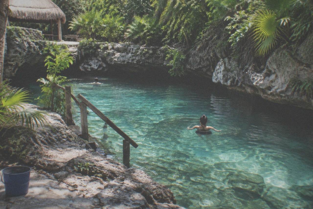 Calm turquoise shallow water in a small open cenote surrounded by limestone rock and green jungle plants