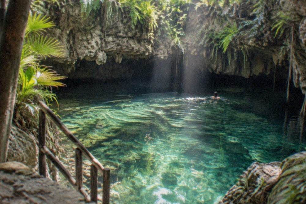 Sunlight beams crossing a cavern cenote opening above clear green water in the late morning