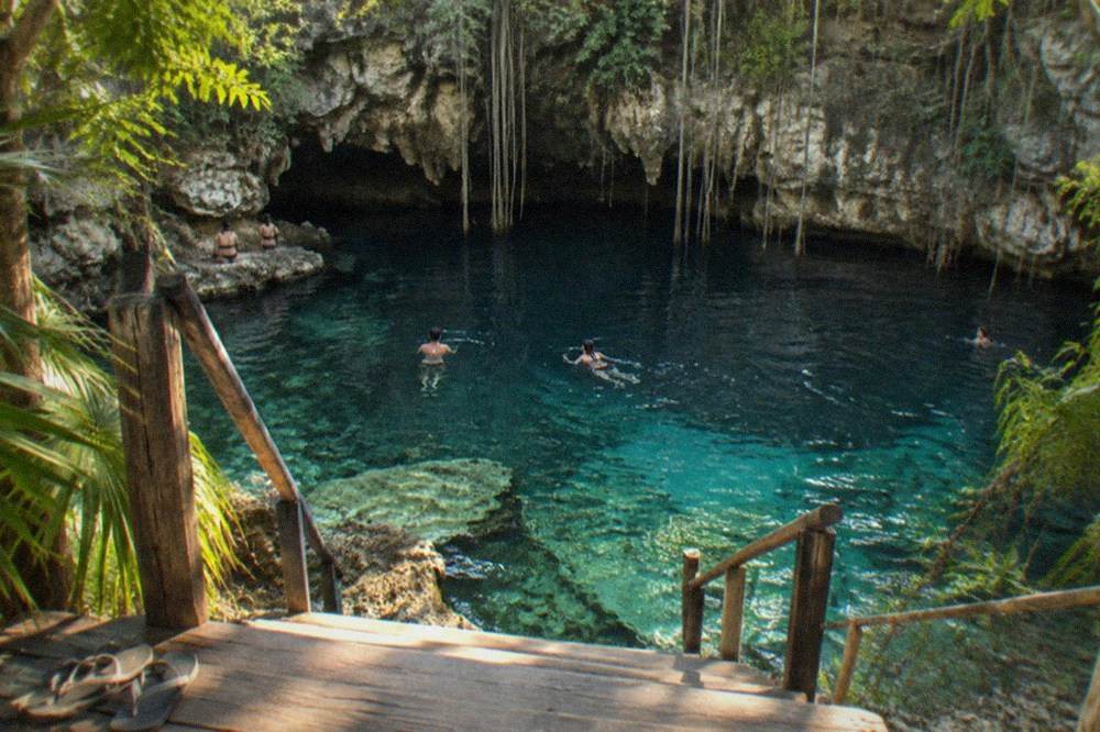 A wooden entry platform and stairs leading down to a clear cenote pool surrounded by limestone rock and hanging vines