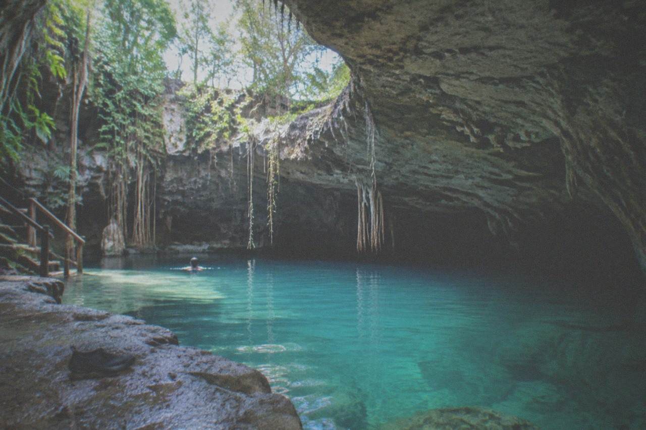 Still turquoise water inside a limestone cenote basin with daylight slanting through an opening in the rock ceiling