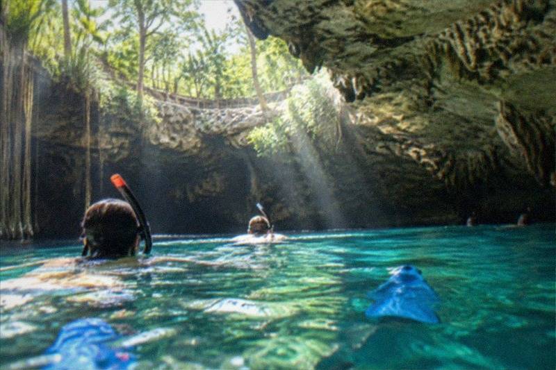 Snorkelers floating over a sunlit Tulum cenote's clear freshwater, light beams entering through the cave mouth