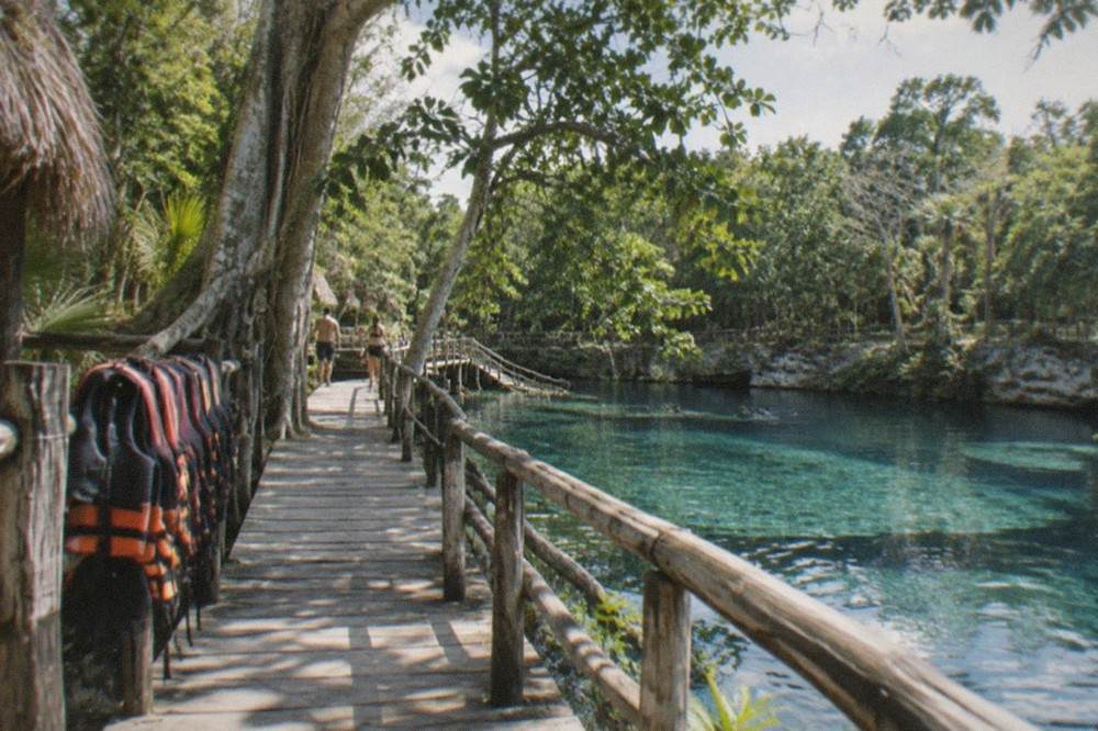 A wide semi-open limestone cenote with pale blue water, wooden walkways and tall jungle trees at the edge