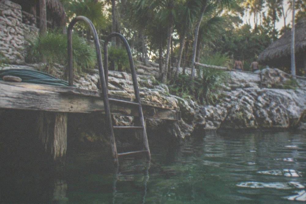 A wooden dock and ladder leading into clear green cenote water surrounded by limestone rock