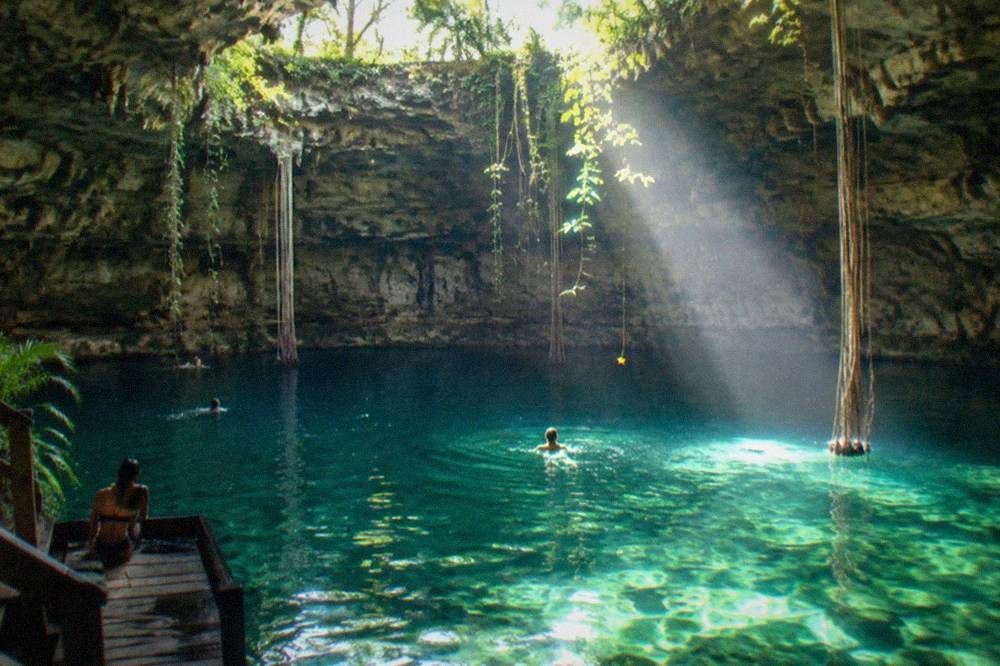 Sunlight beams angling through a round opening in a cave ceiling into green water below