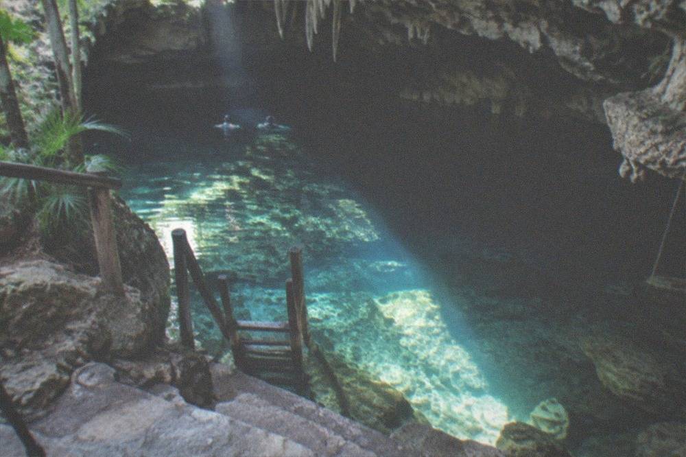 A wooden ladder and stone steps leading down into a dim cavern pool lit by one shaft of light