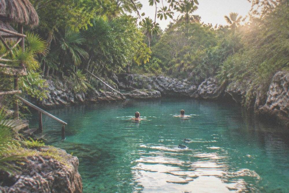 Two swimmers floating on still turquoise water between rock walls and overhanging jungle