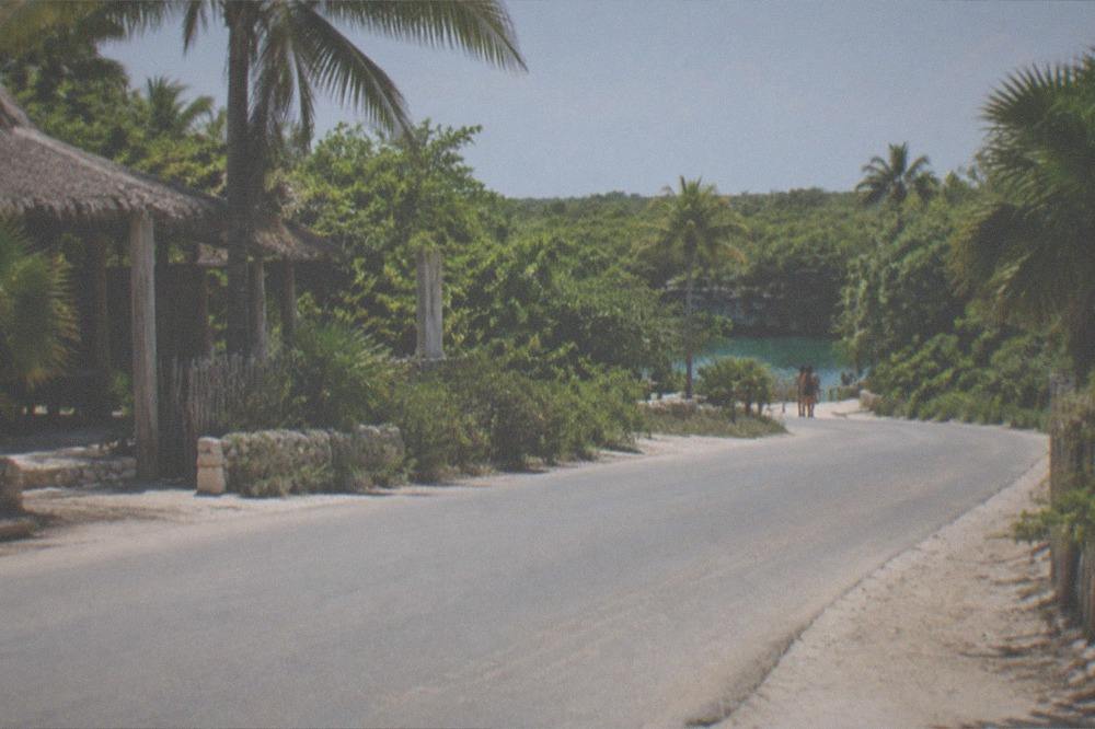 Quiet road curving through low green jungle toward a small cenote entrance sign