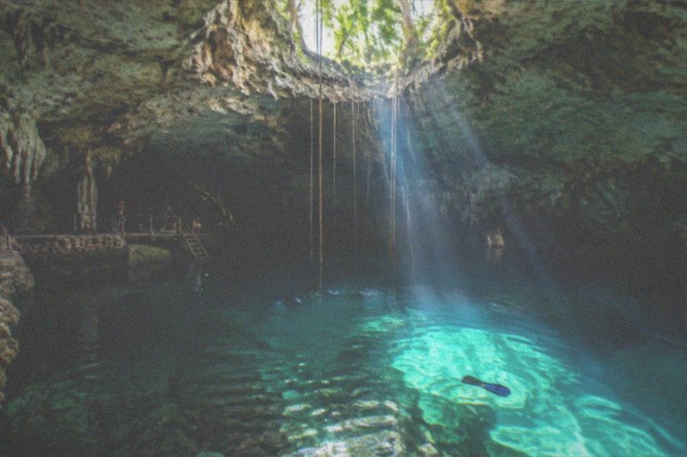 Sunlight beams crossing deep turquoise water inside a limestone cavern opening