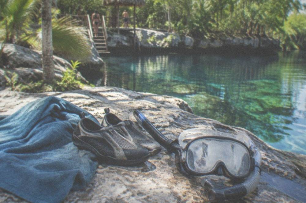 A towel, water shoes and a snorkel mask resting on a rock beside clear cenote water
