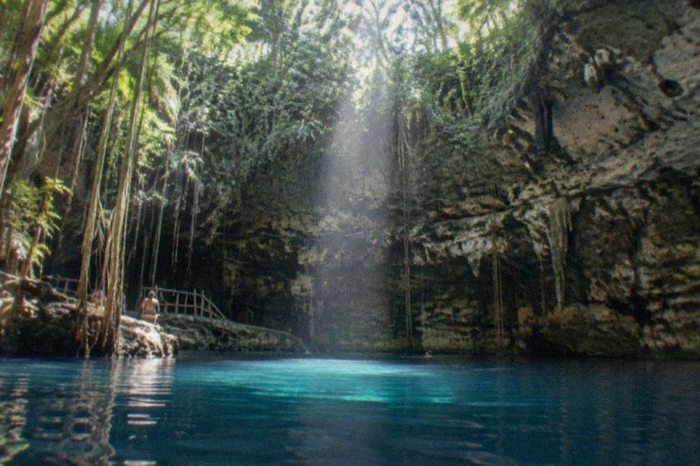 Sunlight beam reaching down through a cenote opening onto still blue water ringed by rock