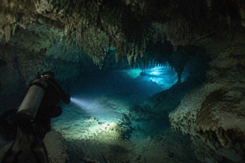 A diver's light illuminating a limestone cavern passage with the blue glow of the entrance visible behind
