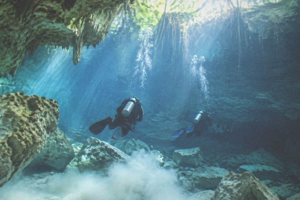 Divers floating motionless in a wide sunlit cavern with underwater rock formations below