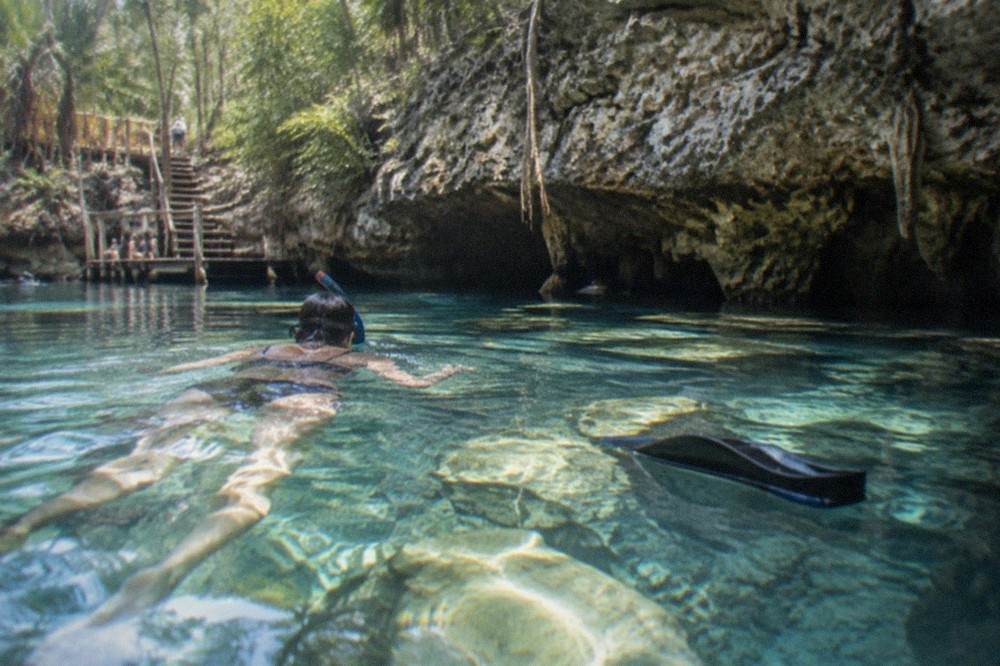 A snorkeler floating face down over pale limestone formations in clear shallow cenote water