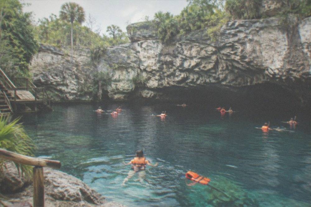 Snorkelers in life vests swimming across a wide open cenote pool beneath limestone walls