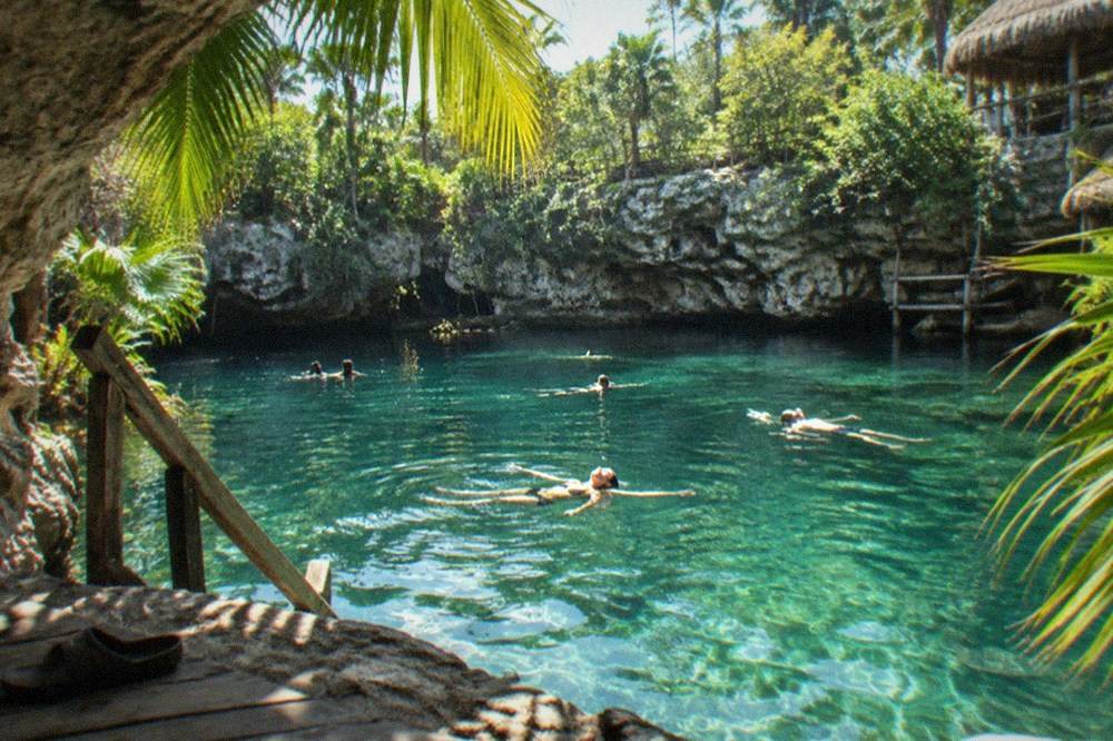 Sunlit circular cenote with a wooden platform and swimmers floating in clear green water