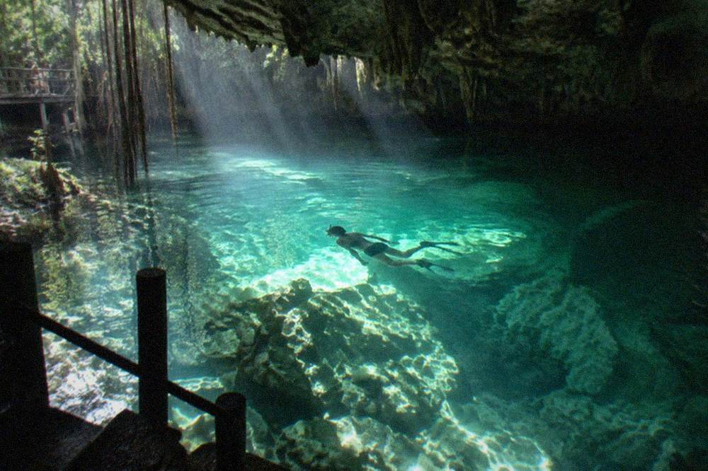 Snorkeler silhouette above pale limestone formations in clear green cave water lit by a shaft of daylight
