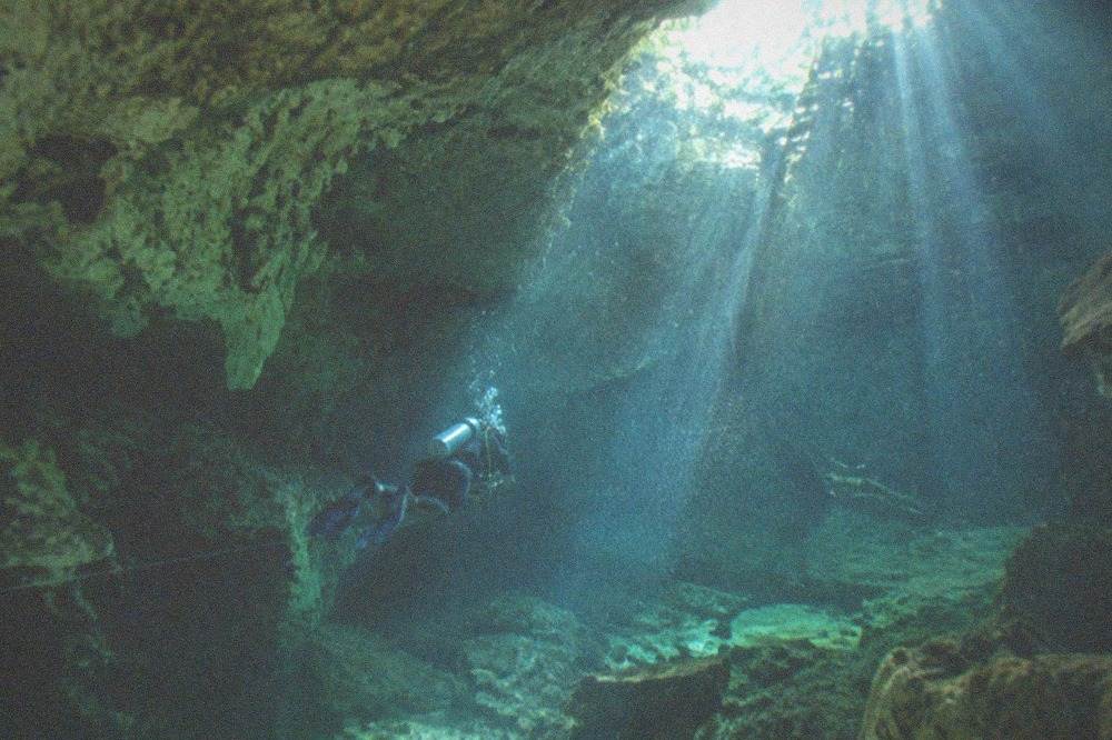Underwater view of a cavern passage with pale rock walls, a guideline and beams of light entering from an opening above