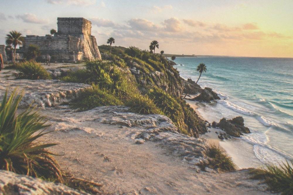 Ancient stone ruin walls on a low cliff above calm turquoise sea and pale sand