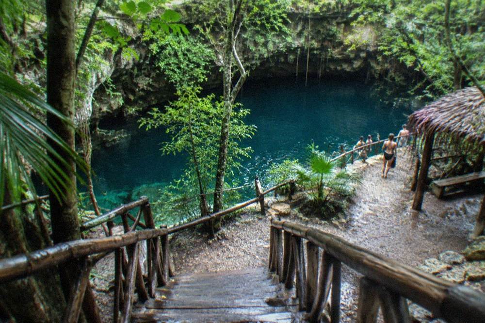 Wooden staircase descending into a round open cenote surrounded by jungle