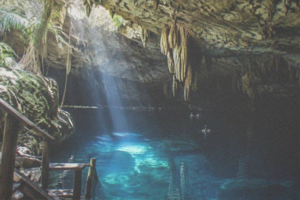 Clear water in a semi-open cenote with stalactites hanging from a low cave ceiling